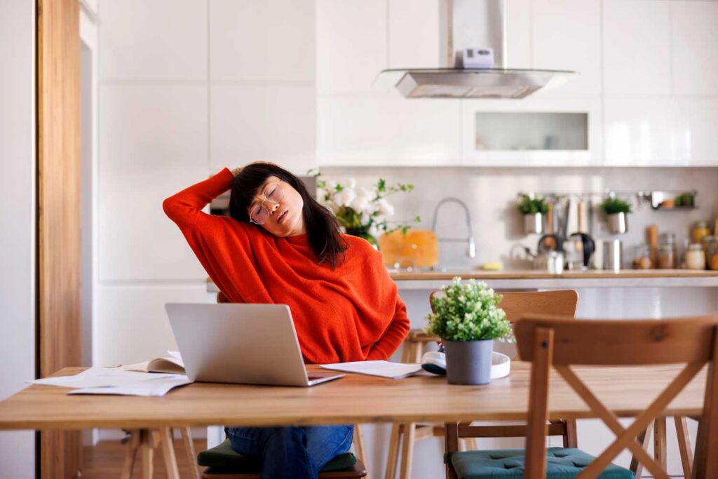 Asian woman sitting at a kitchen table working on a laptop with neck pain